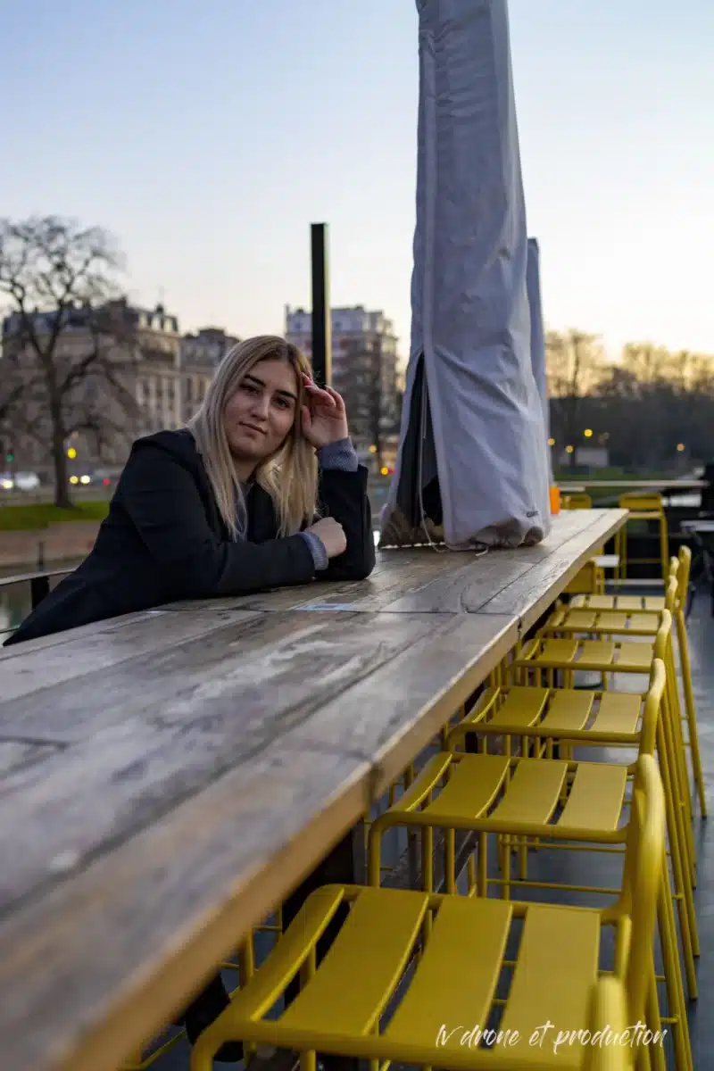 Femme assise à une table en bois avec chaises jaunes, vue sur Lille au coucher du soleil.