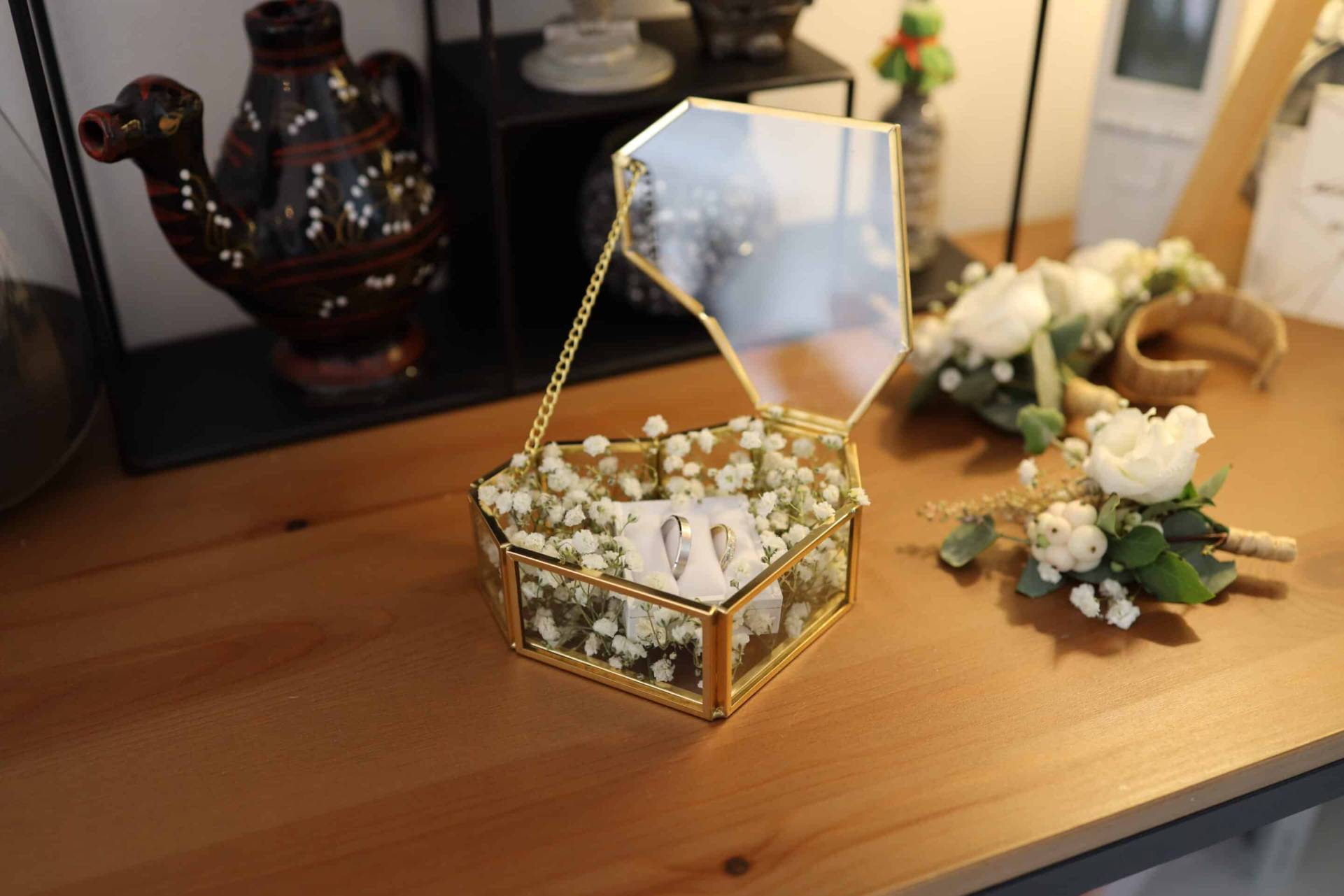 Bague de fiançailles dans une boîte en verre hexagonale avec miroir, décorée de petites fleurs blanc.