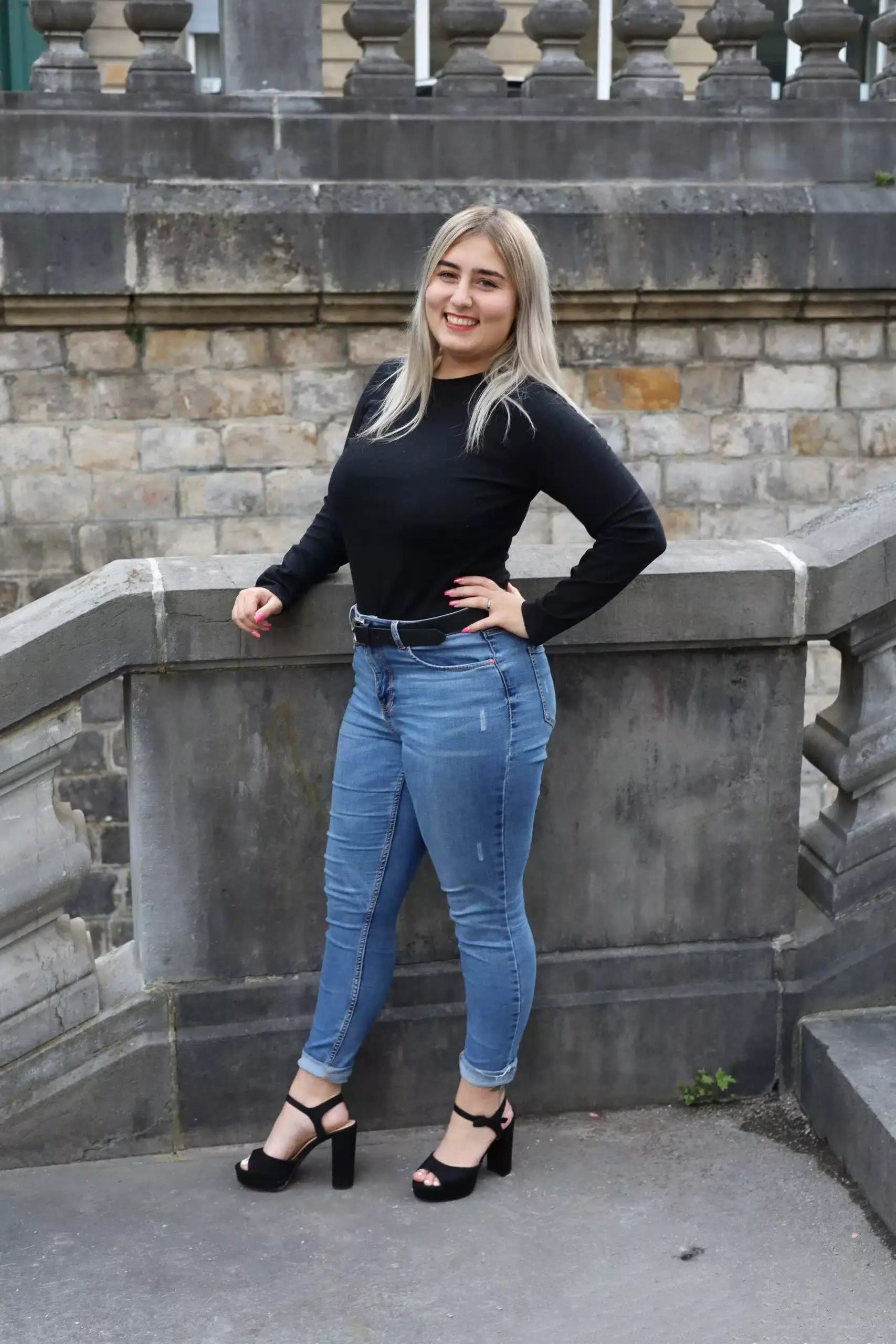 Photographie d'une femme souriante portant un pull noir et un jean bleu, posant devant une structure.