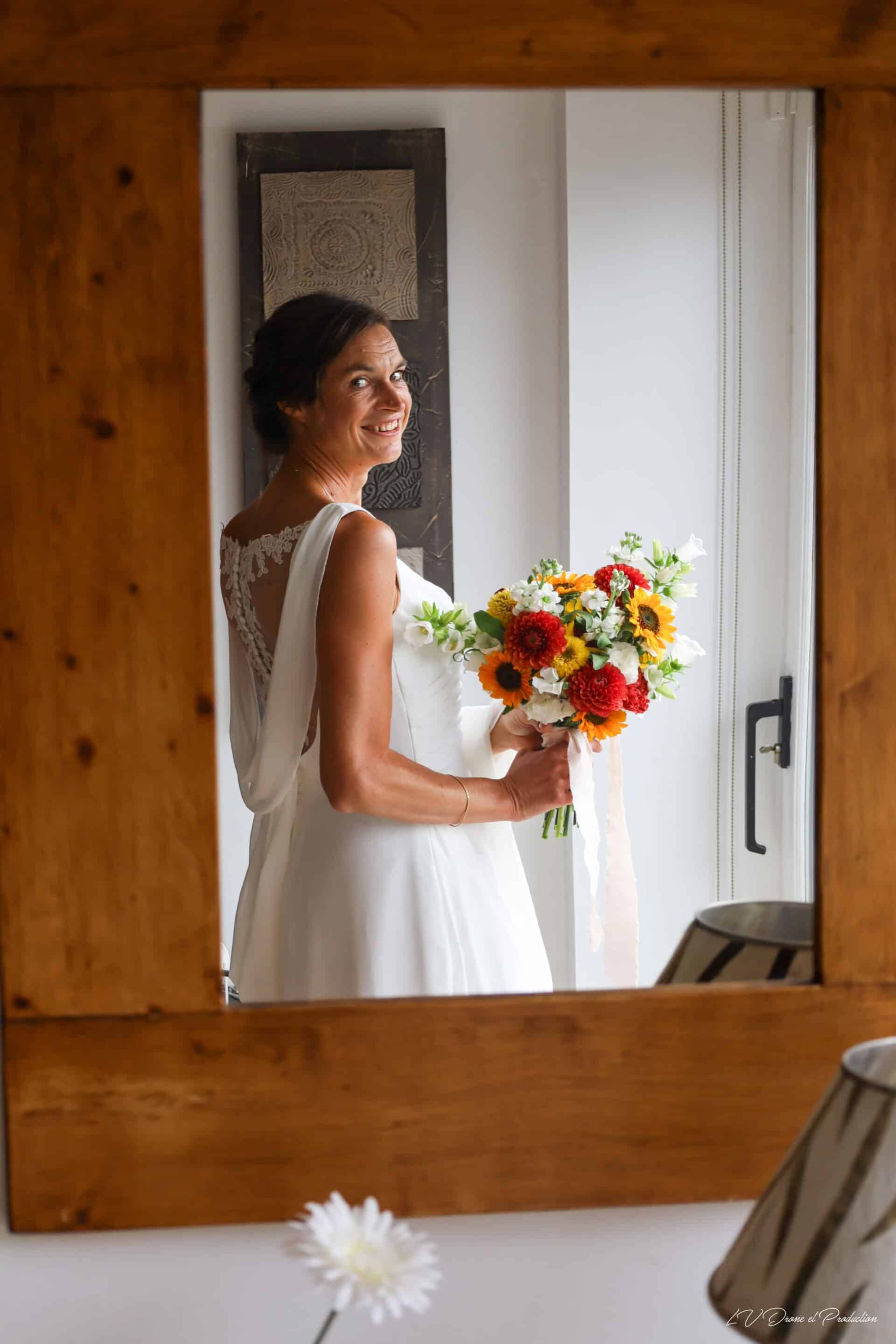 Mariée souriante tenant un bouquet de fleurs colorées dans une pièce lumineuse, reflet dans un miroi.