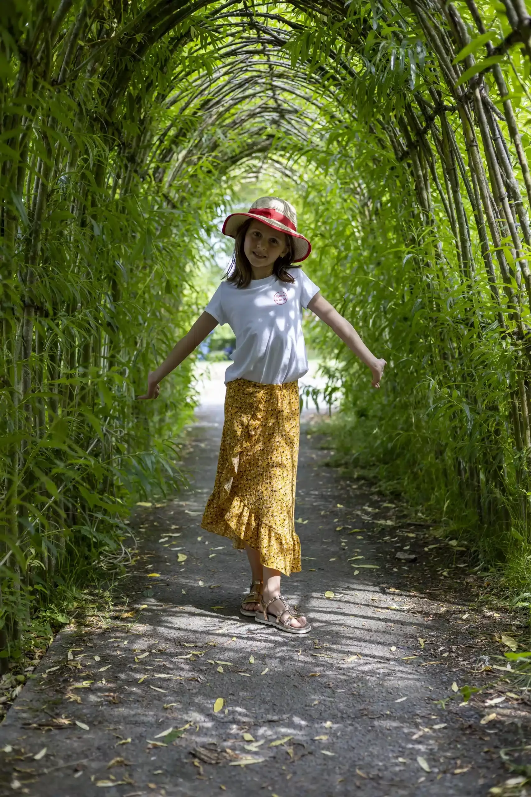 Enfant souriante portant un chapeau rouge dans un tunnel de végétation verte.
